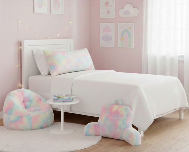 A minimalist girls' bedroom with white bedding featuring a large rainbow pastel shag body pillow, a matching bean bag chair, and a backrest pillow on a white circular rug.